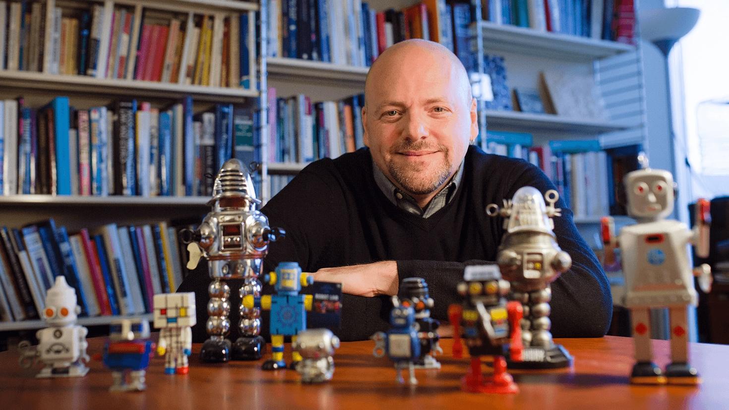 Le professeur Ian Kerr de l’Université d’Ottawa assis à une table où sont disposés des petits robots.
