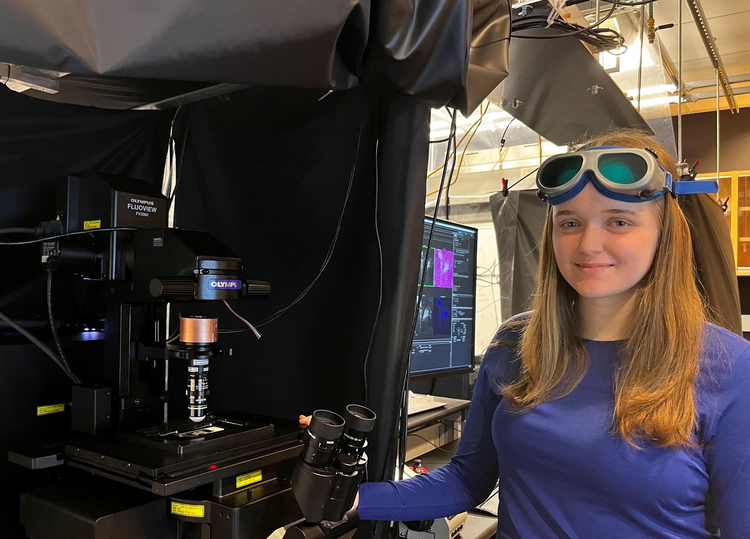 Leah Frackleton is standing next to a laser scanning microscope in her lab. She has goggles on her forehead and one hand holding a microscope