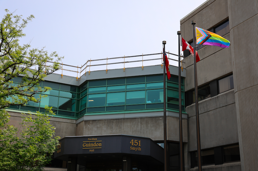 Un drapeau de la fierté flottant aux côtés du drapeau canadien et du drapeau de l’Ontario devant un bâtiment.