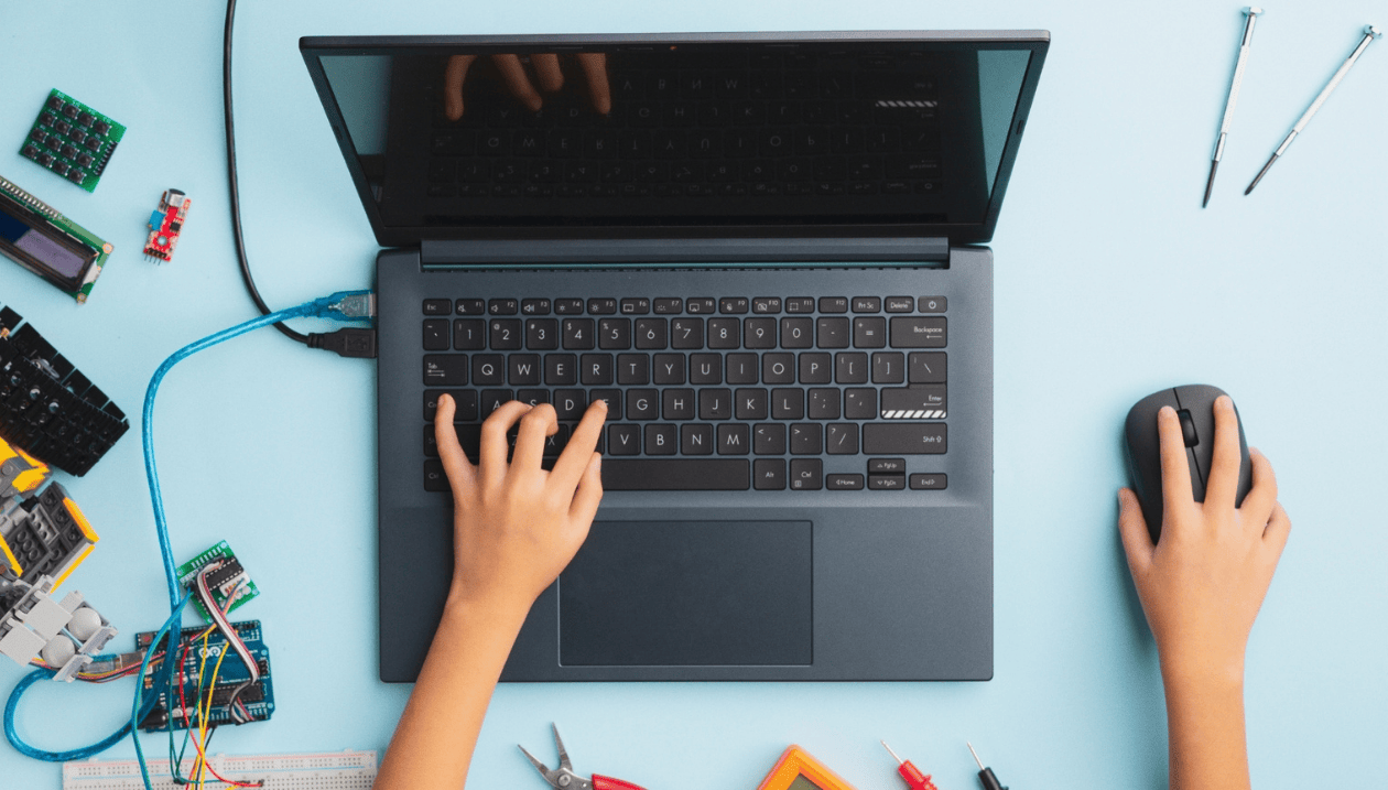 Vue de dessus d’une personne utilisant un ordinateur portable, une main sur le clavier et l’autre sur la souris, entourée de composants électroniques et d’outils sur un espace de travail bleu clair.