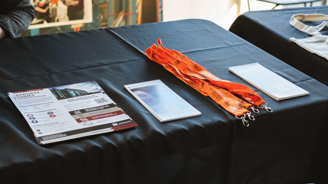 A flat lay shows a black-covered table displaying brochures about graduate studies at the University of Ottawa, a smaller white brochure, and a stack of orange "uOttawa Engineering" lanyards, alongside white pamphlets.