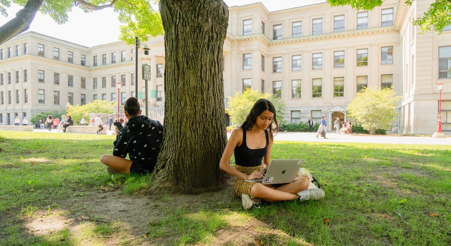 Two students sit under a tree on the Tabaret Lawn.