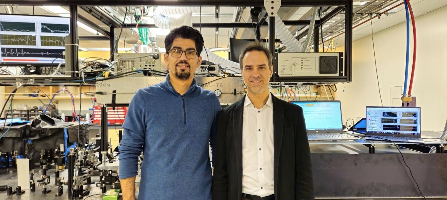 PhD student Ali Maleki (left) and Professor Jean-Michel Ménard (right) standing side by side in a laser laboratory, with complex optical equipment, monitors and wiring visible in the background.