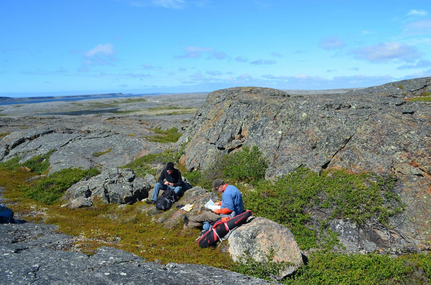 Two researchers, former MSc student Christian Sole (left) and Professor Jonathan O’Neil (right), sit among large rock outcrops while conducting geological fieldwork on the Nuvvuagittuq Greenstone Belt in Nunavik, northern Quebec.
