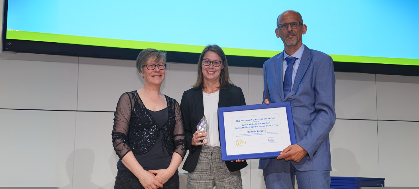 (From left to right) Marian Holness (vice-president of the European Geosciences Union), Renelle Dubosq and Peter van der Beek (president of the European Geosciences Union) standing together on stage. Renelle Dubosq is holding a crystal-shaped trophy, and Peter van der Beek is holding a framed certificate dedicated to Renelle for the Arne Richter Award for Outstanding Early Career Scientists.