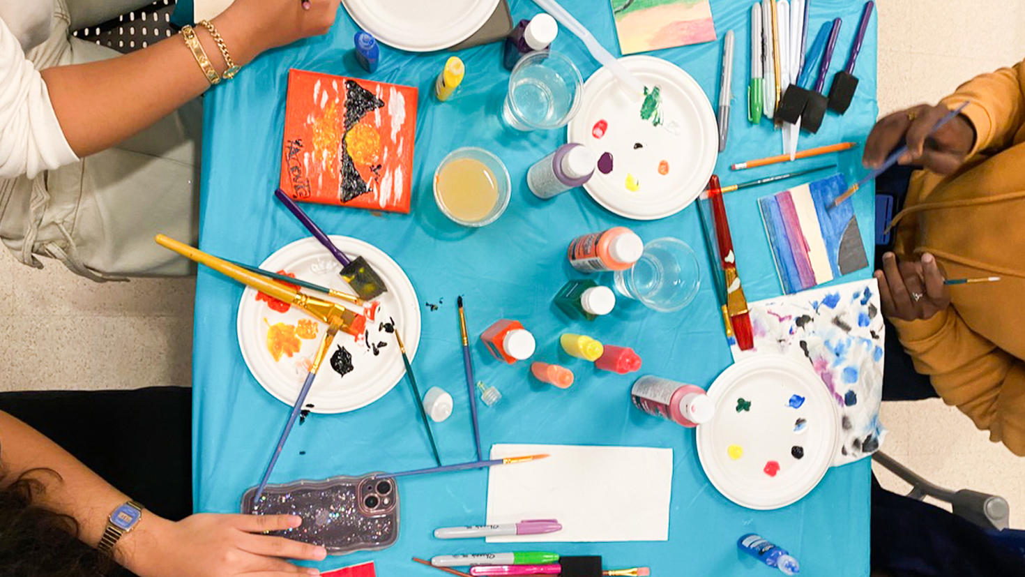 An overhead view of scattered paintbrushes, paint, markers and other art materials on a table.