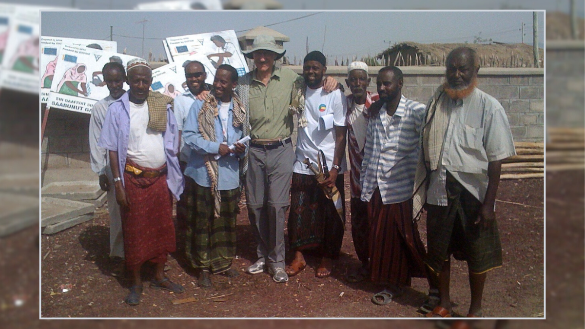 A group of nine men stands outside, some are smiling and have their arms around each other. Most of the men are wearing button down shirts with sarongs, but the man in the middle is wearing pants.