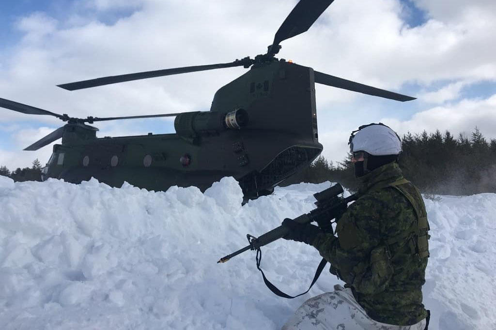 Une personne debout dans la neige avec une arme, devant un hélicoptère militaire.