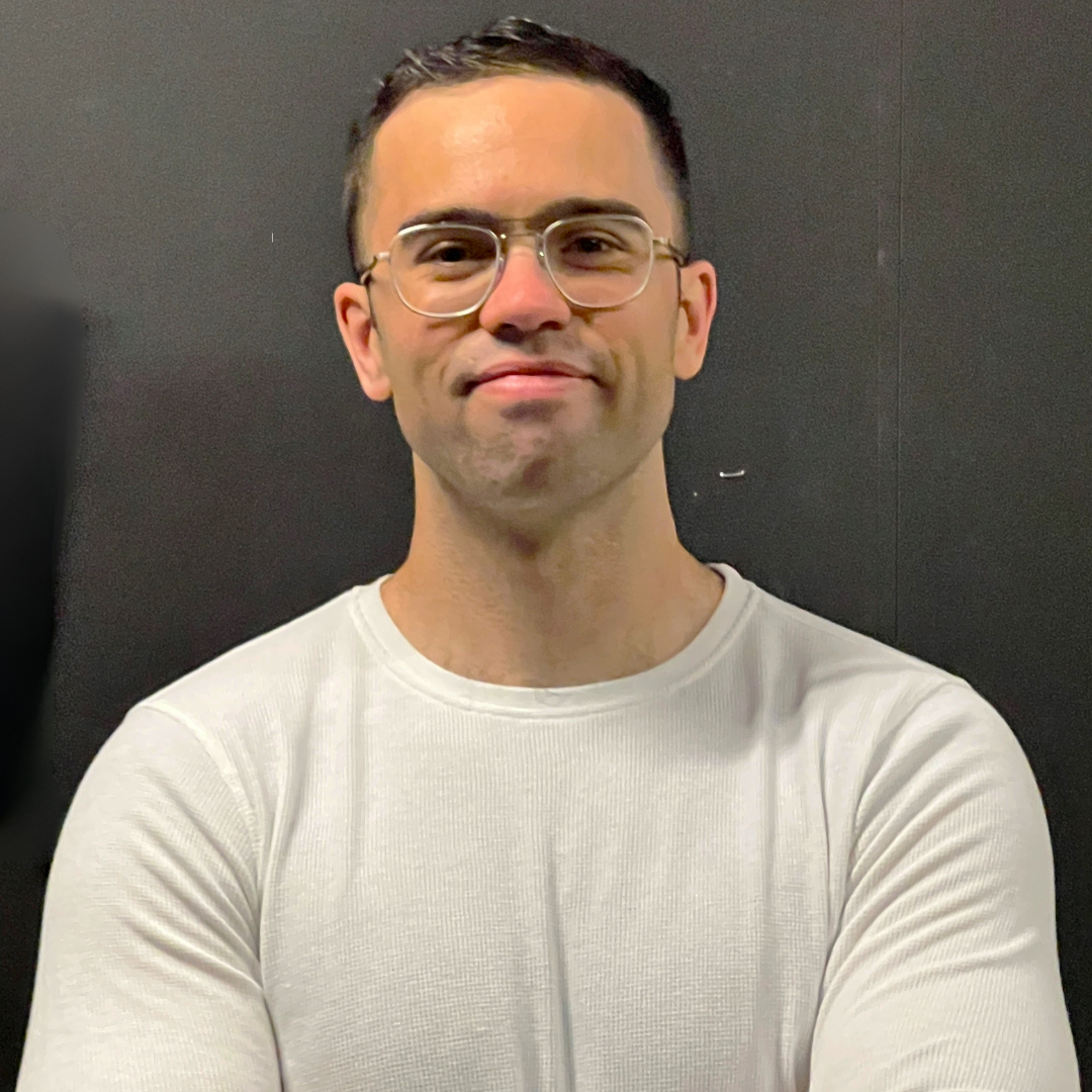 Simon Larose pose les bras croisés devant un fond sombre, vêtu d’un chandail blanc à manches longues et portant des lunettes.