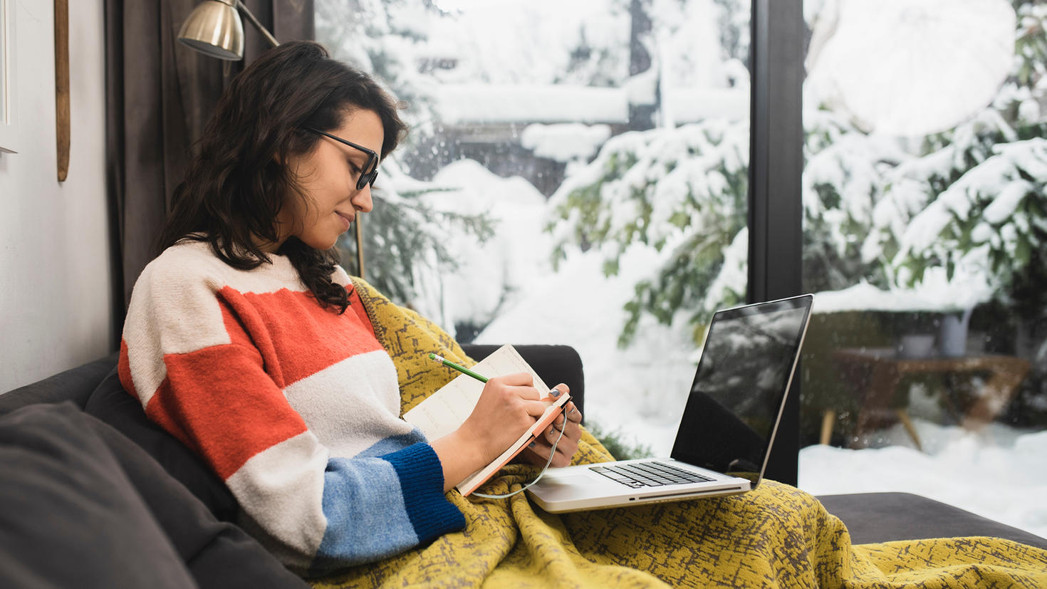 A woman wrapped under a blanket studies on a sofa, taking notes.
