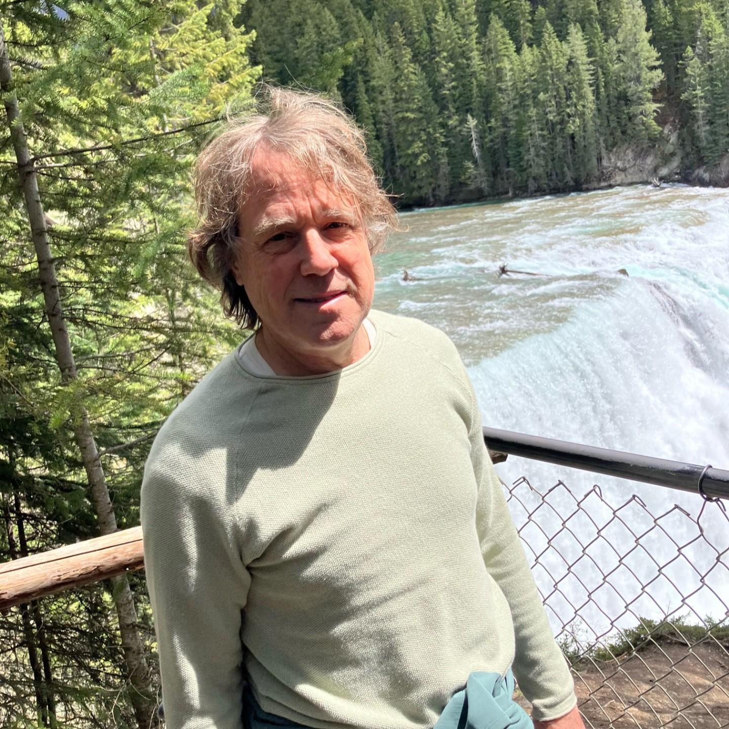 Professor André Longtin stands near a waterfall surrounded by trees.