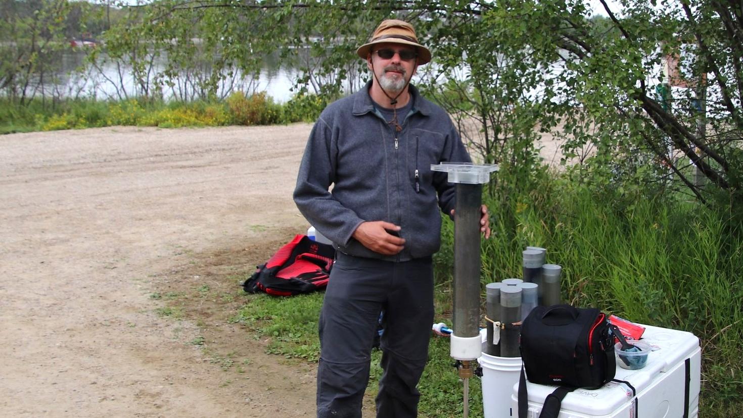 Professor Jules Blais stands outdoors near a lakeshore, wearing field gear and a wide-brimmed hat. He’s holding a long sediment core sample tube and standing beside scientific sampling equipment, including coolers and containers.