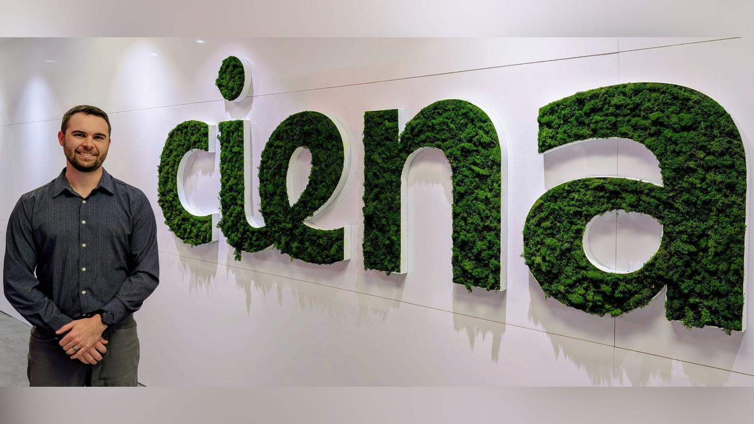 Alumnus Nicolas Couture stands, smiling, beside a large, green Ciena logo.