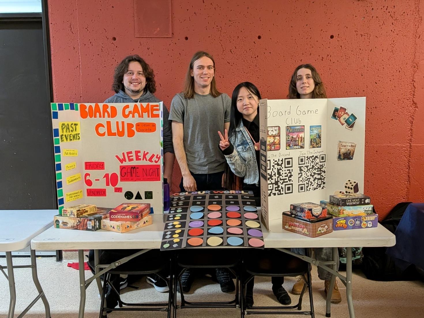 Les dirigeants du Club de jeux de société de l'Université d'Ottawa debout derrière leur table à la foire des clubs d'hiver, avec deux présentoirs à trois volets, des piles de jeux de société et le jeu personnalisé du club, Colour Wordle, au centre.
