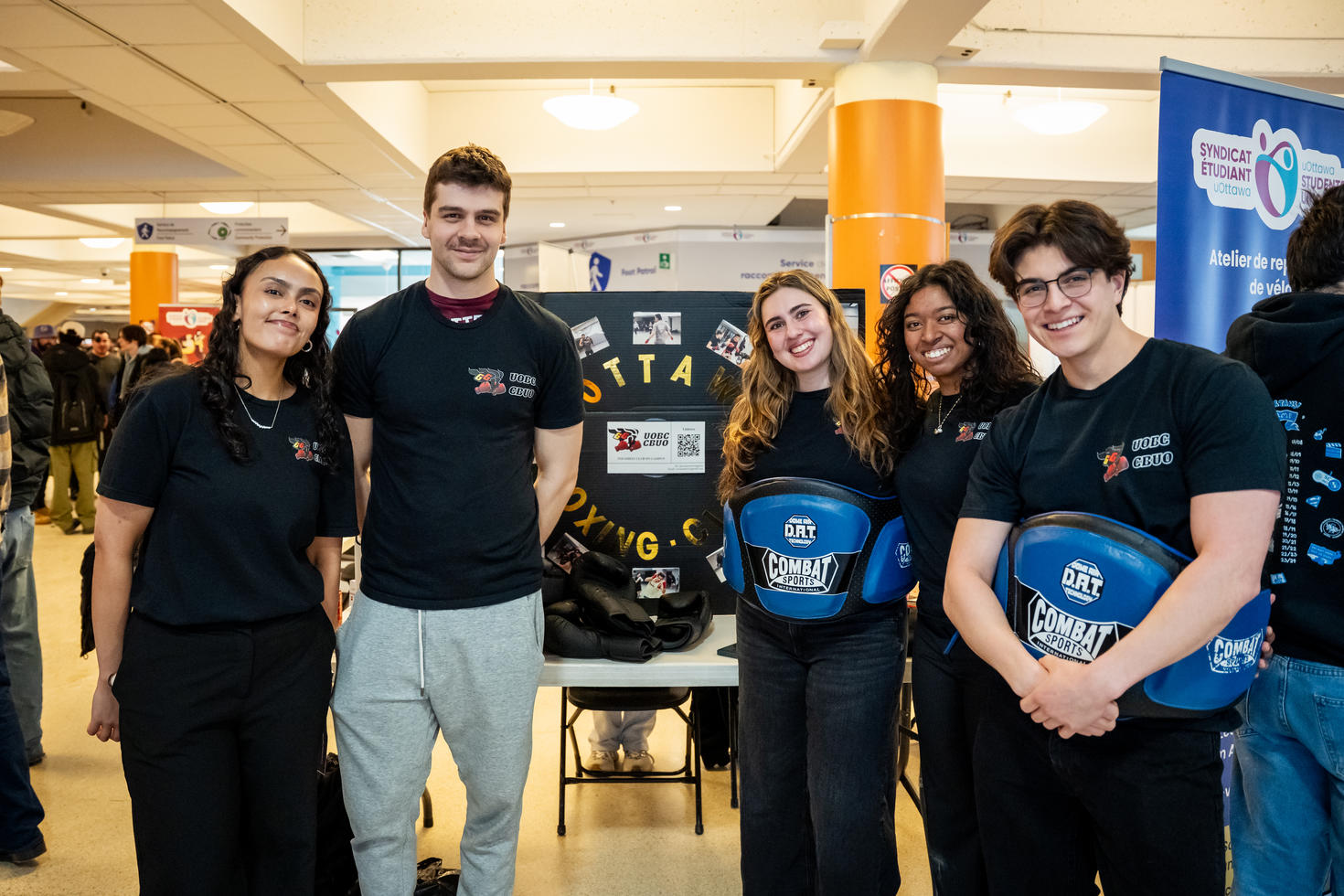 Les dirigeants du Club de boxe de l'Université d'Ottawa devant le stand de leur club à la Foire des clubs d'hiver. Deux d'entre eux portent un équipement de protection bleu autour de la taille. Derrière eux, un panneau noir à trois volets affiche le nom du club en lettres dorées, un logo, un code QR et des photos de boxe.