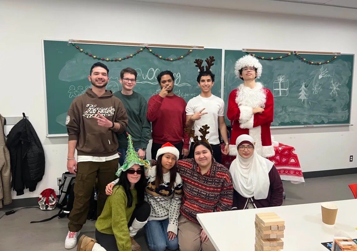 Les dirigeants du club de cuisine de l'Université d'Ottawa en costumes de fête/de Noël posant devant un tableau noir dans une salle de classe après un événement.