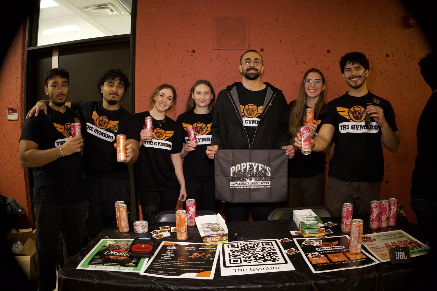 Les dirigeants de GymBro à la foire des clubs d'hiver, debout derrière une table recouverte de boissons énergisantes GURU, d'un code QR, d'autocollants, de barres et de dépliants. Les sept membres portent des t-shirts noirs assortis avec le logo du club et tiennent des canettes de boissons énergisantes, tandis que le membre au centre tient une bannière Popeye's Supplements.