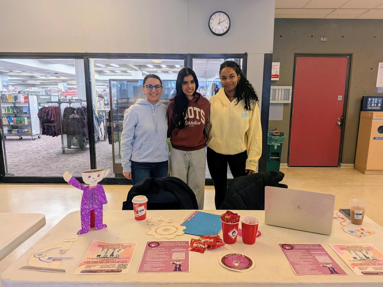 Les dirigeants de Pipetting Hot Tea à la foire des clubs d'hiver, debout derrière une table avec des affiches, des bonbons, un ordinateur portable et des découpes en papier en forme d'engrenages, un microscope, leur logo et un personnage agitant la main avec une tasse de thé à la place de la tête.