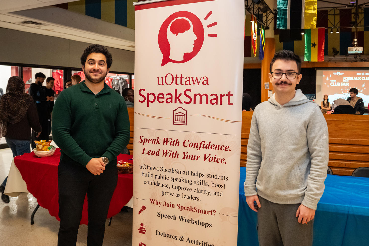 Les dirigeants du club SpeakSmart de l'Université d'Ottawa debout de chaque côté d'une bannière lors du salon des clubs d'hiver. La bannière porte l'inscription « Speak With Confidence. Lead With Your Voice. » (Parlez avec confiance. Dirigez avec votre voix.) et met en avant la mission et les activités du club.