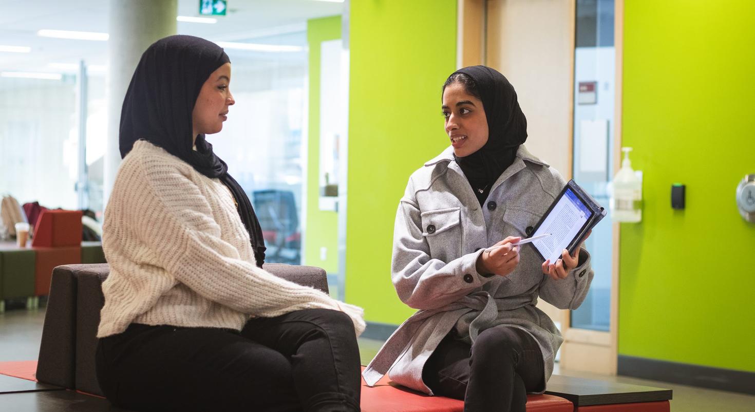 A student holding a tablet points to the screen as she talks to another student.