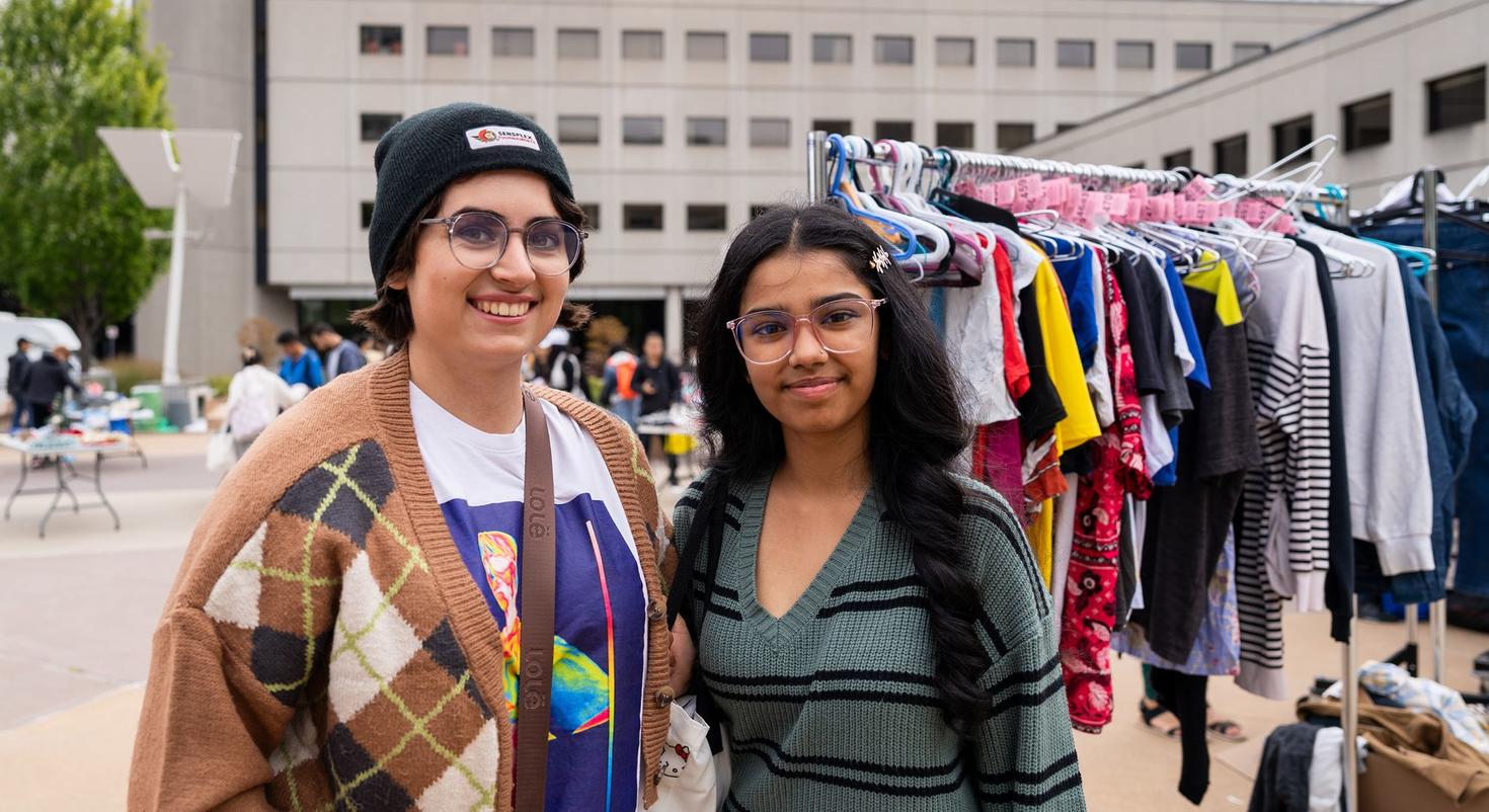 Deux personnes souriant à la caméra lors d'une Gratuiterie éphémère , debout devant des portants remplis de vêtements donnés.