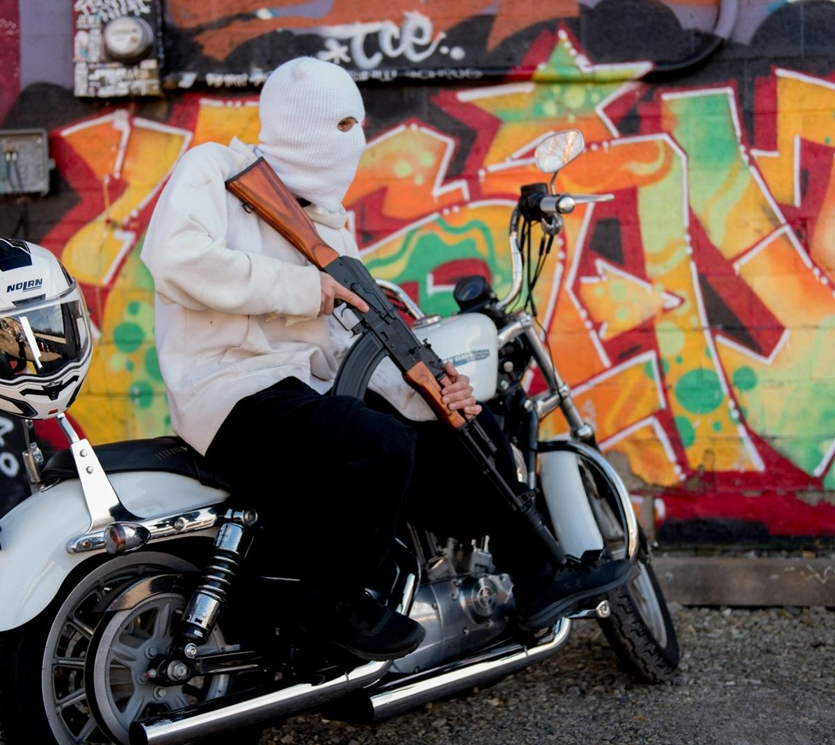 Un homme cagoulé tient un AK silencieux sur une moto devant un graffiti coloré.