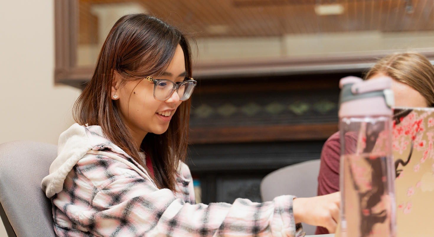 Une étudiante souriante portant des lunettes travaille avec enthousiasme sur son ordinateur portable dans un espace d'étude du campus.