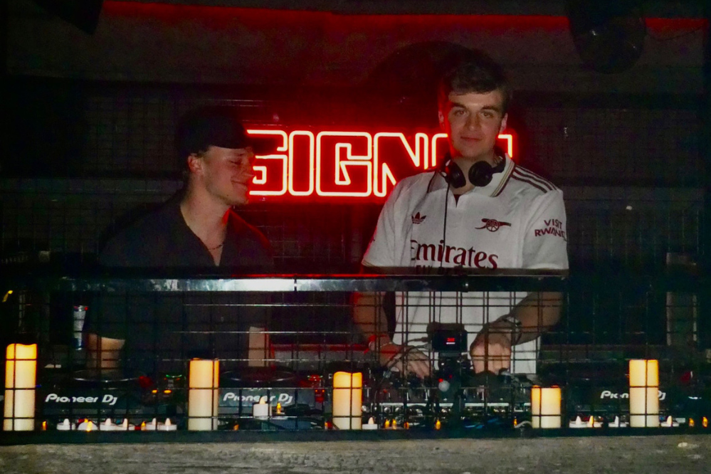 Deux jeunes hommes se tiennent derrière une table de DJ. L'un d'eux sourit à la caméra.