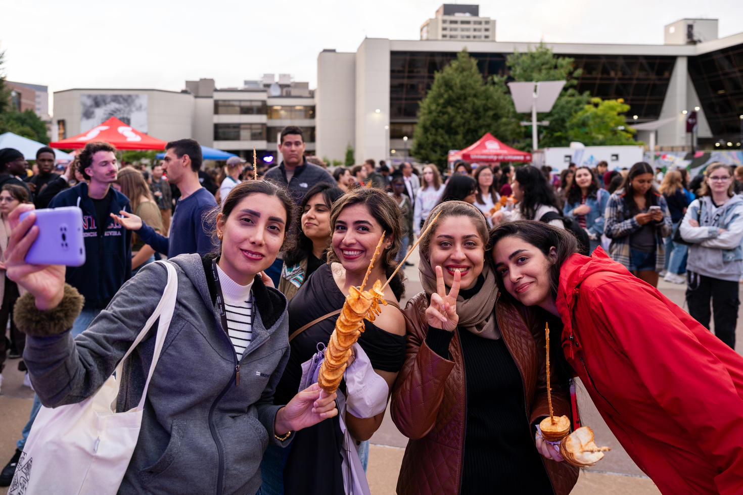 Four students smile for a selfie while enjoying food at a busy outdoor campus event, with a crowd gathered in the background. 