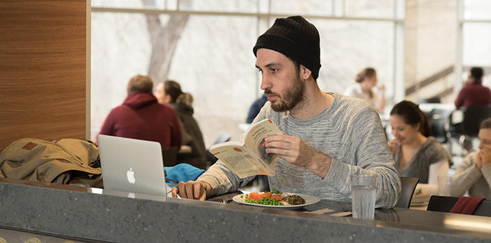 A man wearing a beanie and holding a book uses his laptop while eating.