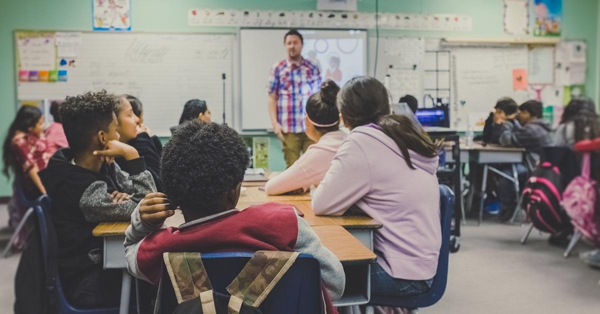 : Vus de dos, des élèves assis dans une salle de classe écoutent leur enseignant.