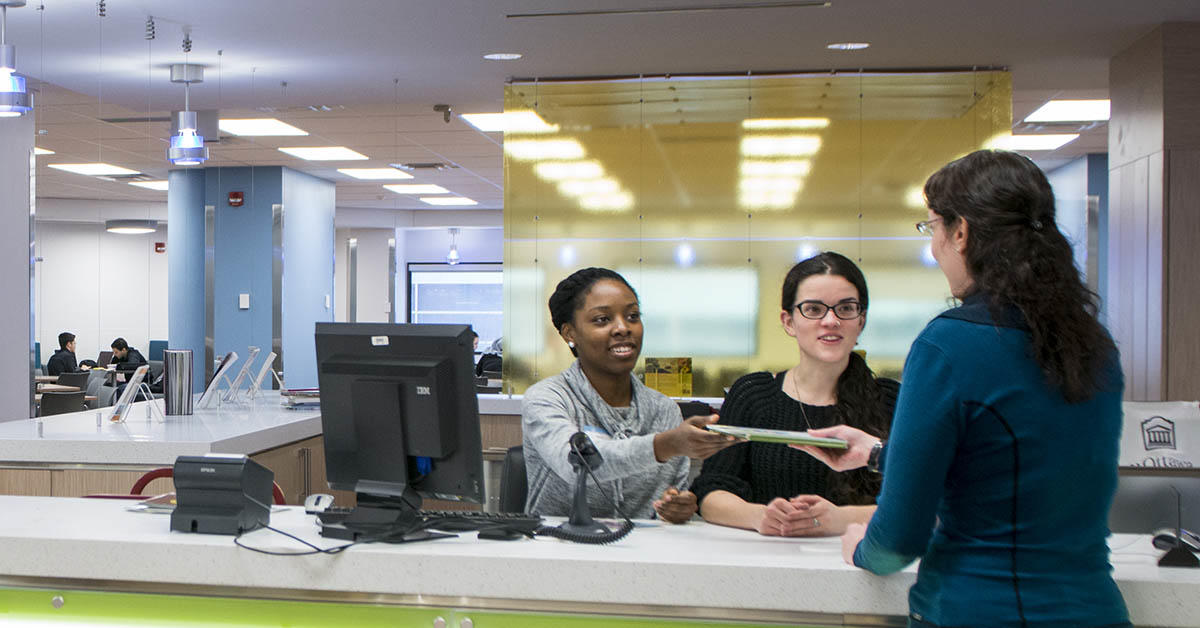 Une employée de la bibliothèque, accompagnée d’une collègue, tend un document à une étudiante qui se tient devant un comptoir.