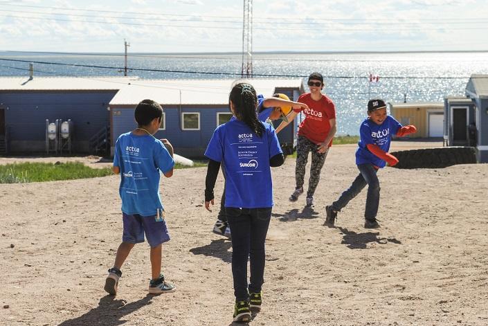 Children running around an open area with a few houses, a lake and an older guide in the background.