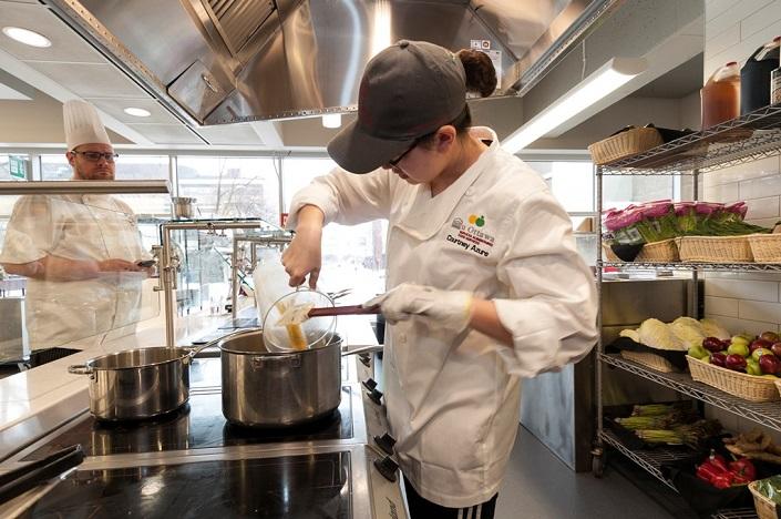 Woman pouring sauce into a cauldron as a chef looks on.