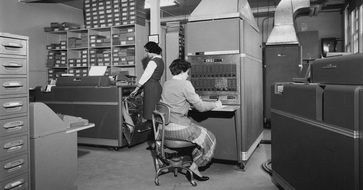 Une femme est assise à un ordinateur de première génération alors qu’une autre se tient à côté.