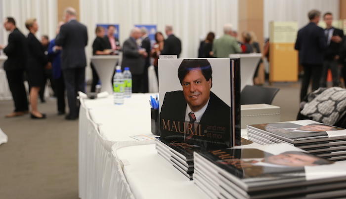 Dans un salon où des gens discutent à l’arrière-plan, plusieurs exemplaires du même livre sont posés par piles sur une table. L’un d’eux, placé debout, montre une photo of Mauril Bélanger.