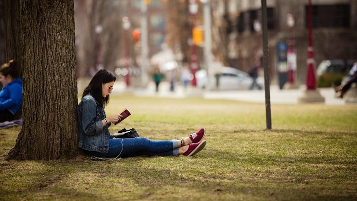 Une étudiante lit un livre, assise contre un arbre sur la pelouse devant le pavillon Tabaret.