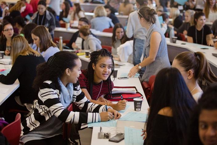 Quatre jeunes femmes conversent en classe, d’autres font de même en arrière-plan. 