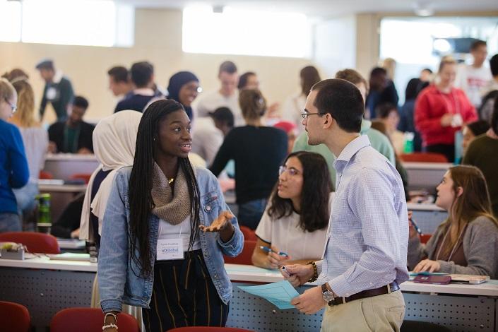 Deux étudiants discutent en classe; derrière eux, un groupe diversifié d’étudiants est aussi en discussion. 