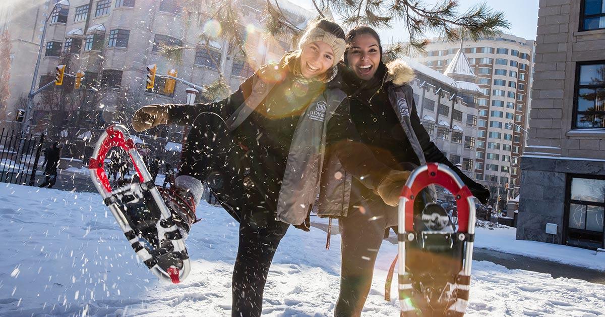 Two students, each smiling and lifting a leg up in the snow to display a snowshoe.