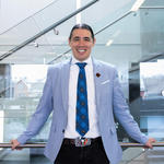 Portrait du professeur Robert-Falcon Ouellette, portant un blazer bleu clair, se tenant devant un fond en verre.